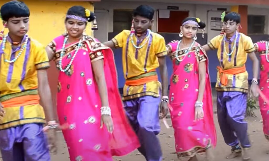 A group of men and women performing the Dhimsa dance, forming a circle and moving in synchronized steps. The dancers clap their hands and stamp their feet in rhythm with the music